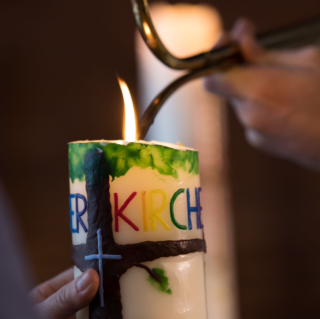 Nahaufnahme einer brennenden Kerze. Auf der Kerze sind ein braunes Kreuz und bunte Buchstaben. Man erkennt die Buchstaben E R links und K I R C H rechts vom Kreuz.