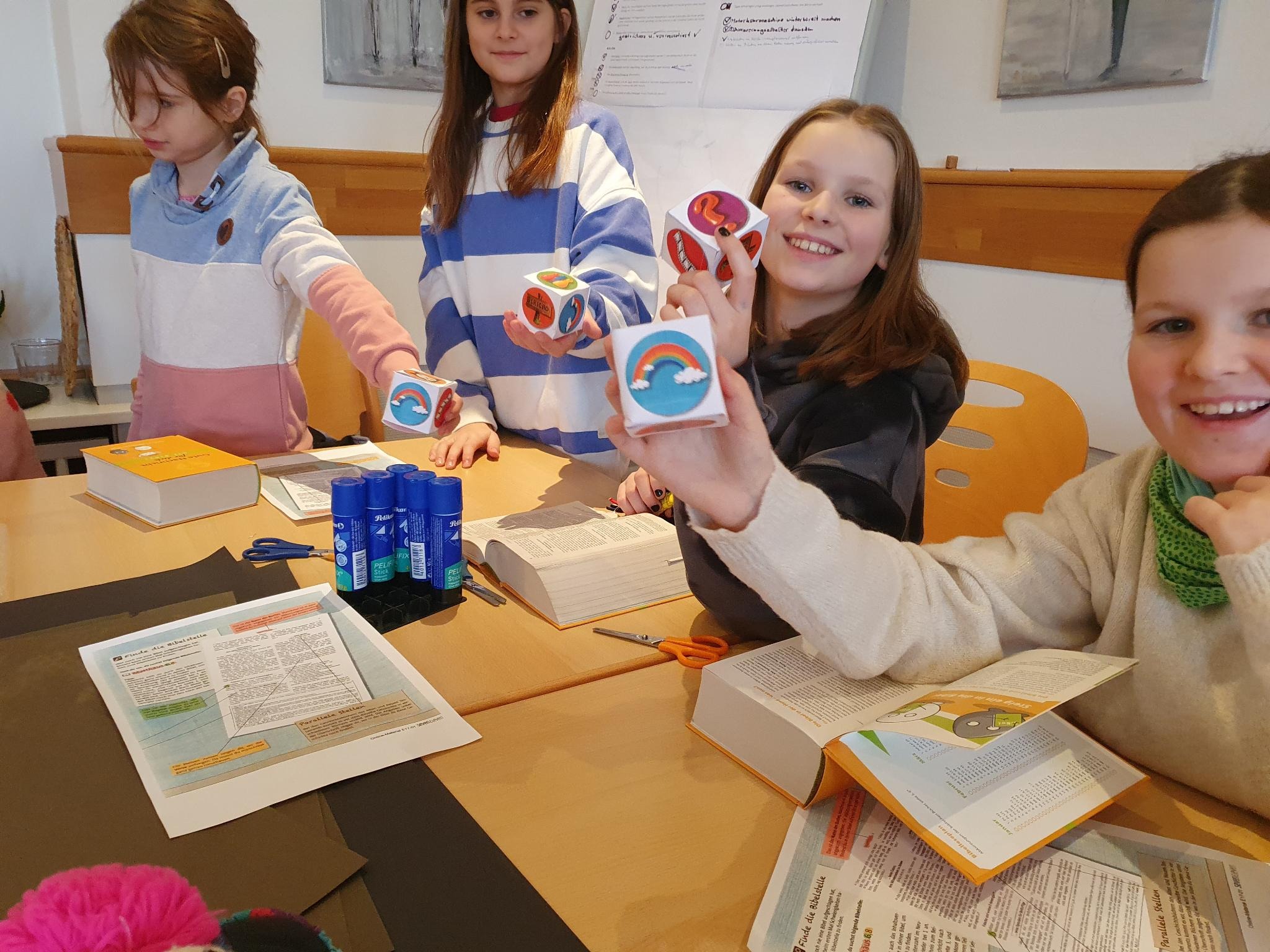 Vier Kinder sitzen an einem Basteltisch und zeigen lachend ihre bemalten Würfel in die Kamera.