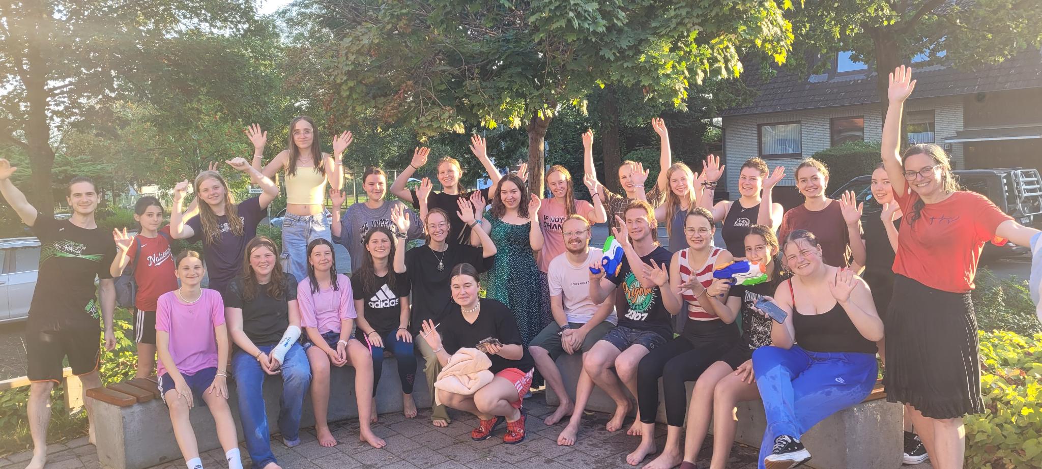 Bild einer Jugendgruppe. Es ist offenbar Sommer, weil alle Jugendlichen kurze Hosen und T-Shirts tragen. Im Hintergrund sind Bäume zu sehen.